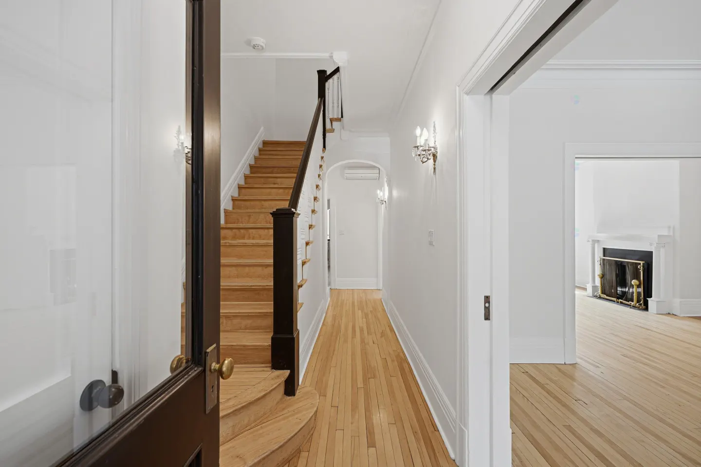 Bright hallway with hardwood floors, white walls, and a wooden staircase with a dark brown banister. A fireplace is visible through a doorway.