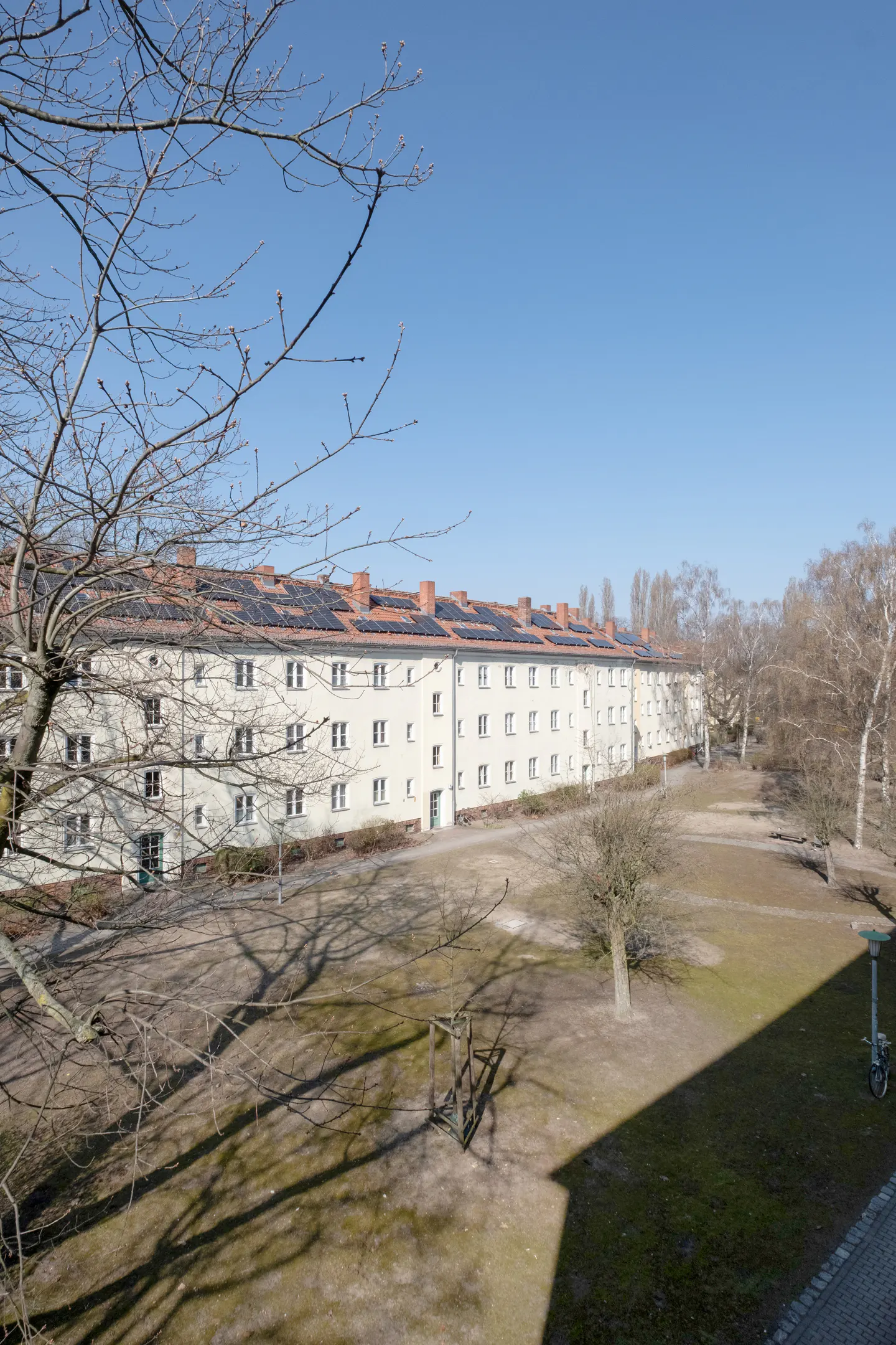 View of a long, light-yellow apartment building with solar panels on the roof, facing a grassy courtyard with bare trees under a clear blue sky.