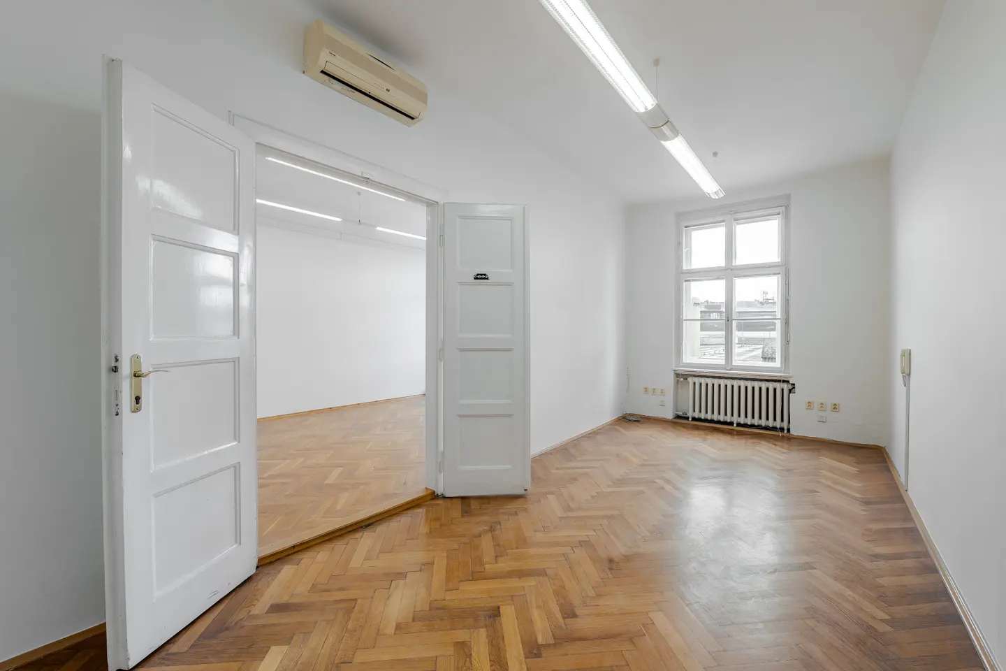 Bright, empty room with herringbone wood floors, white walls, and open white doors to another room. A window lets in natural light.