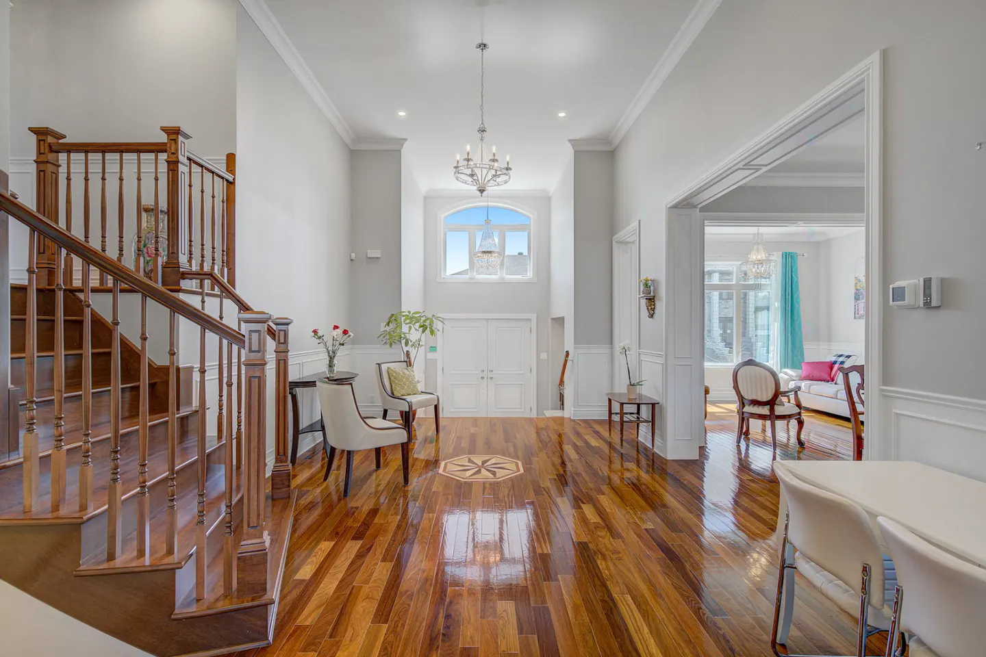 Bright foyer with hardwood floors, white walls, and a staircase on the left. A chandelier hangs above the front door. A living room is visible through an archway.