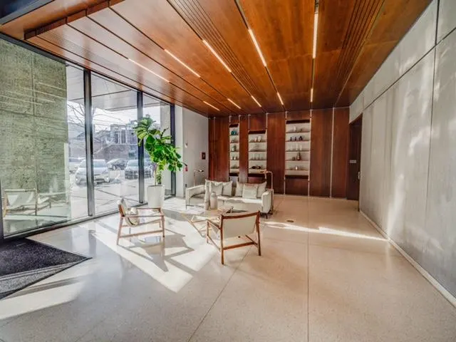 Modern lobby with wood ceiling, large windows, and seating area. A potted plant sits near the entrance. Shelves line the back wall.