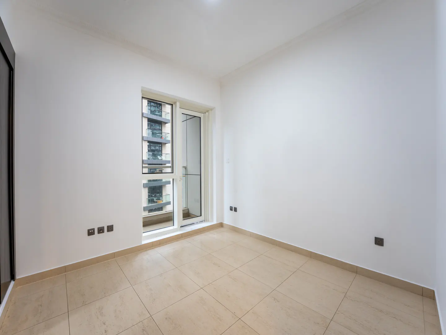 Bright, empty room with beige tile floor, white walls, and a sliding glass door to a balcony with a view of a high-rise building.
