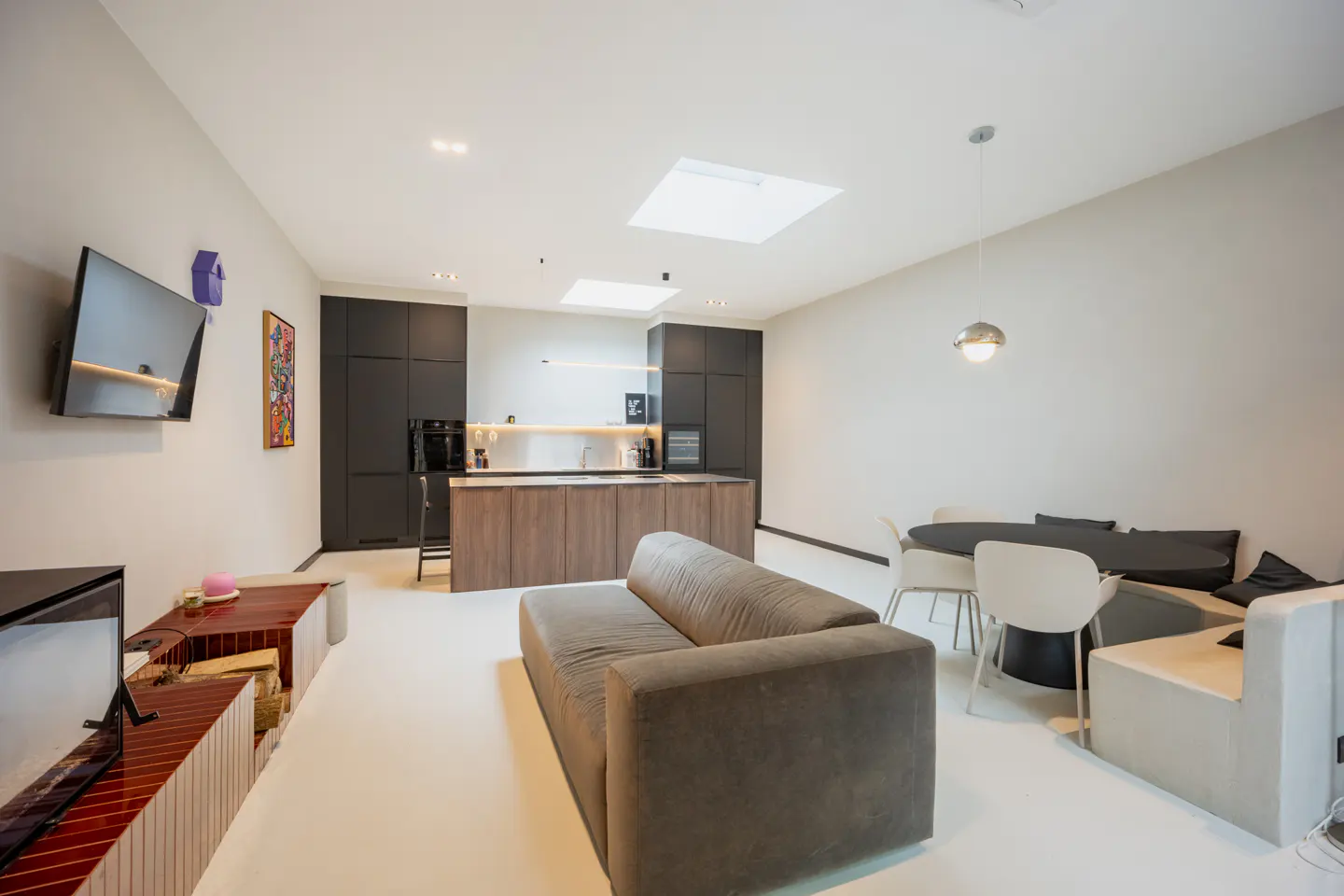 A modern, open-concept living space with a gray sofa, black kitchen cabinets, and a round dining table. Skylights provide natural light.