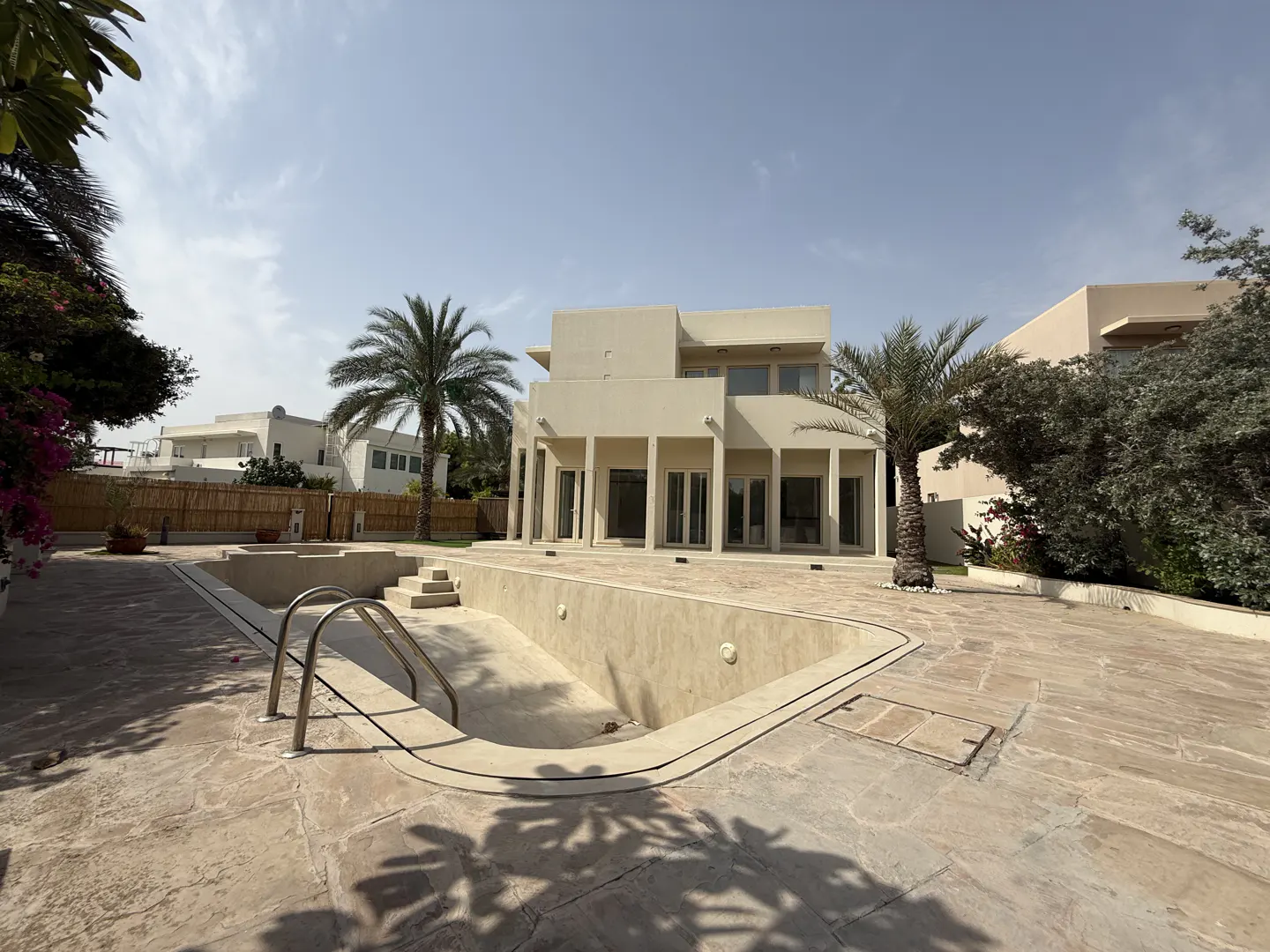 Exterior view of a beige two-story house with an empty pool in the foreground and palm trees in the yard.