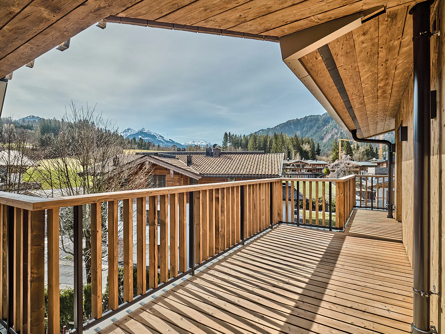 View from a wooden balcony with a wooden railing, overlooking houses, trees, and mountains in the distance.