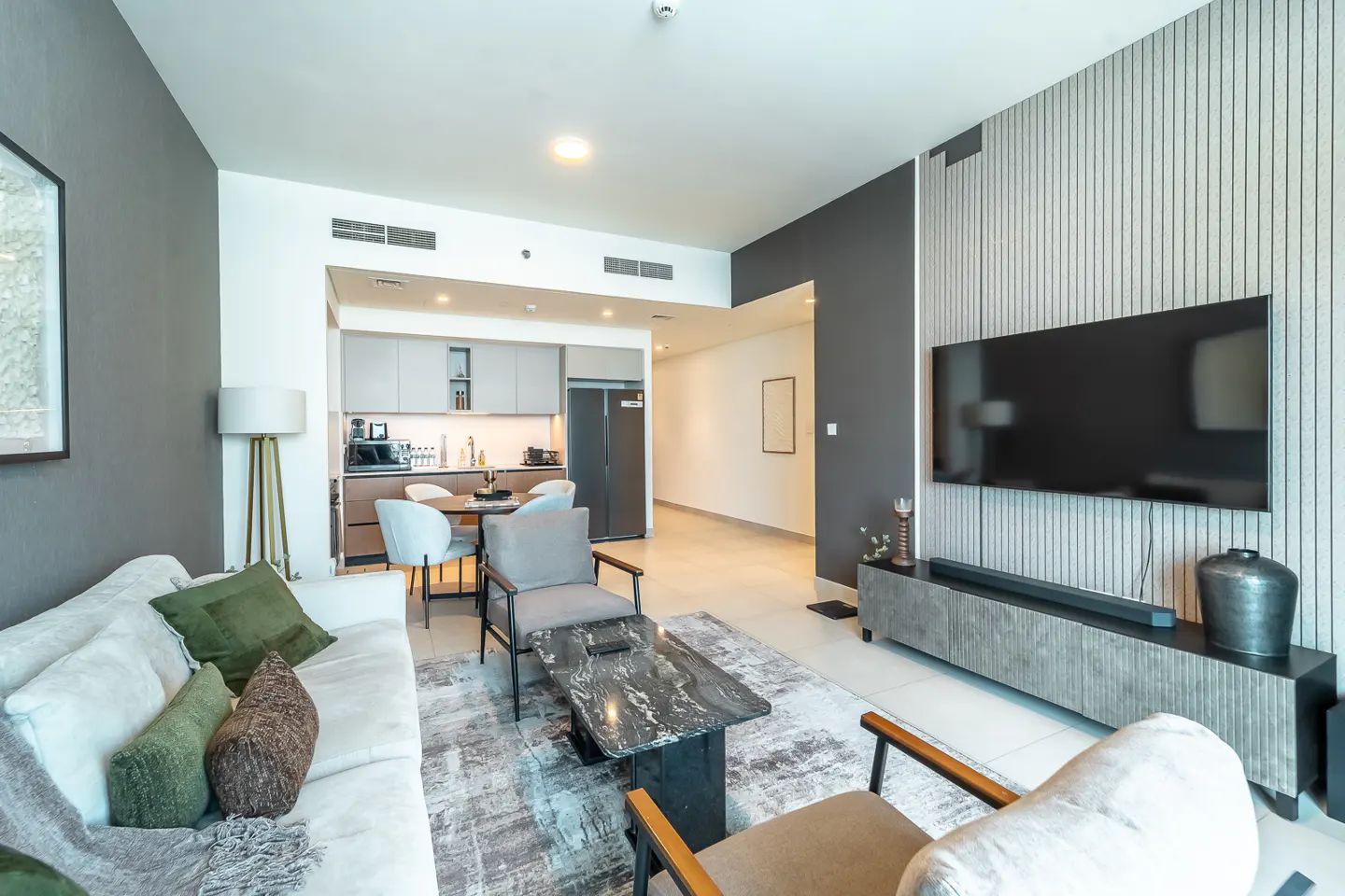 Modern apartment living room with white sofa, gray chairs, marble table, and open kitchen in the background.