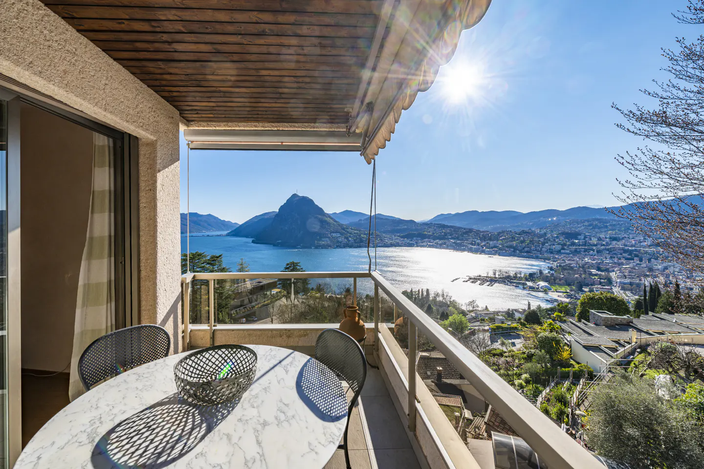 Balcony view of Lake Lugano, Switzerland. A marble table and chairs sit on the balcony overlooking the lake and mountains on a sunny day.