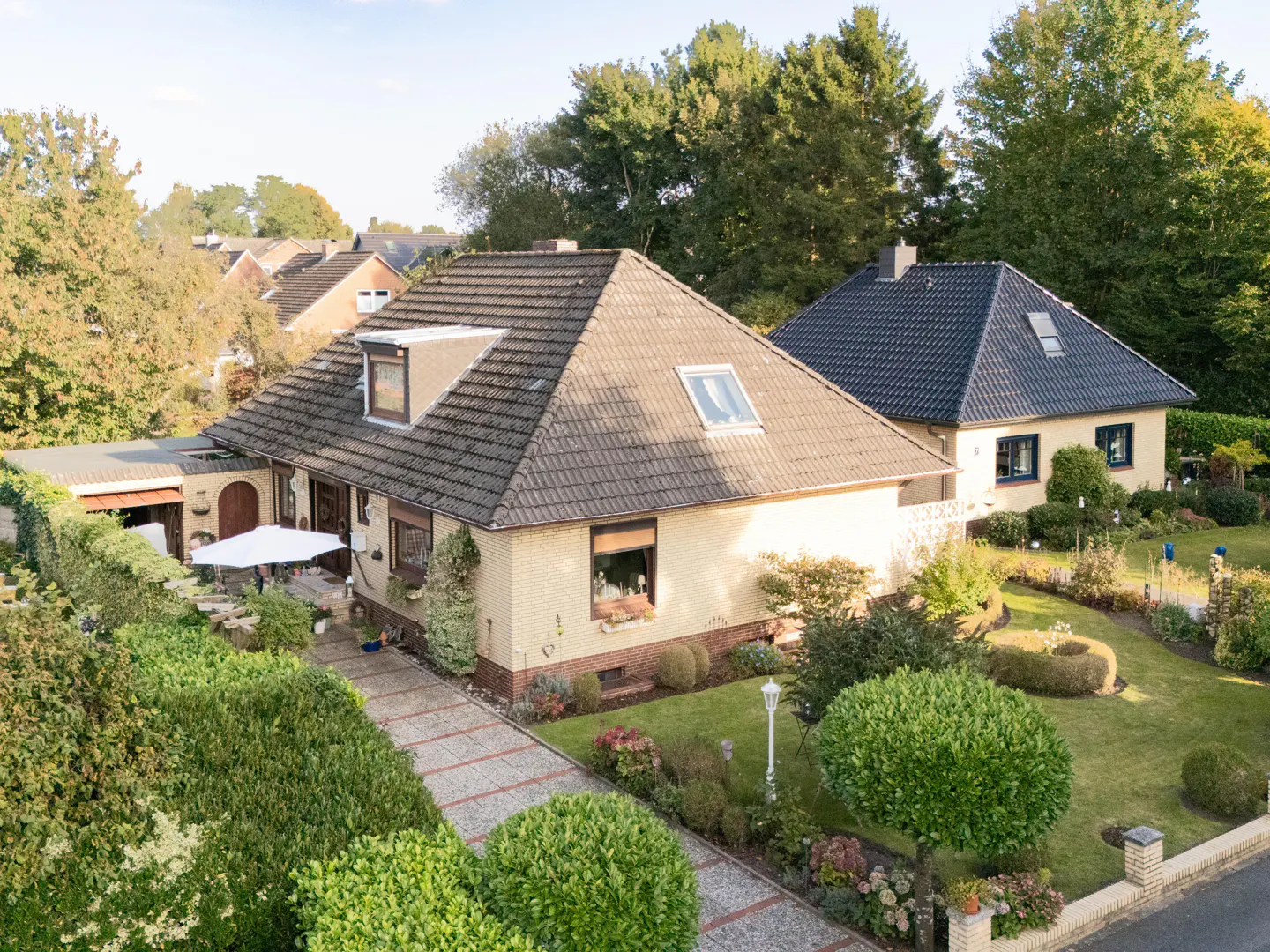 Aerial view of a yellow brick house with a brown tiled roof, surrounded by a lush green garden and trees.