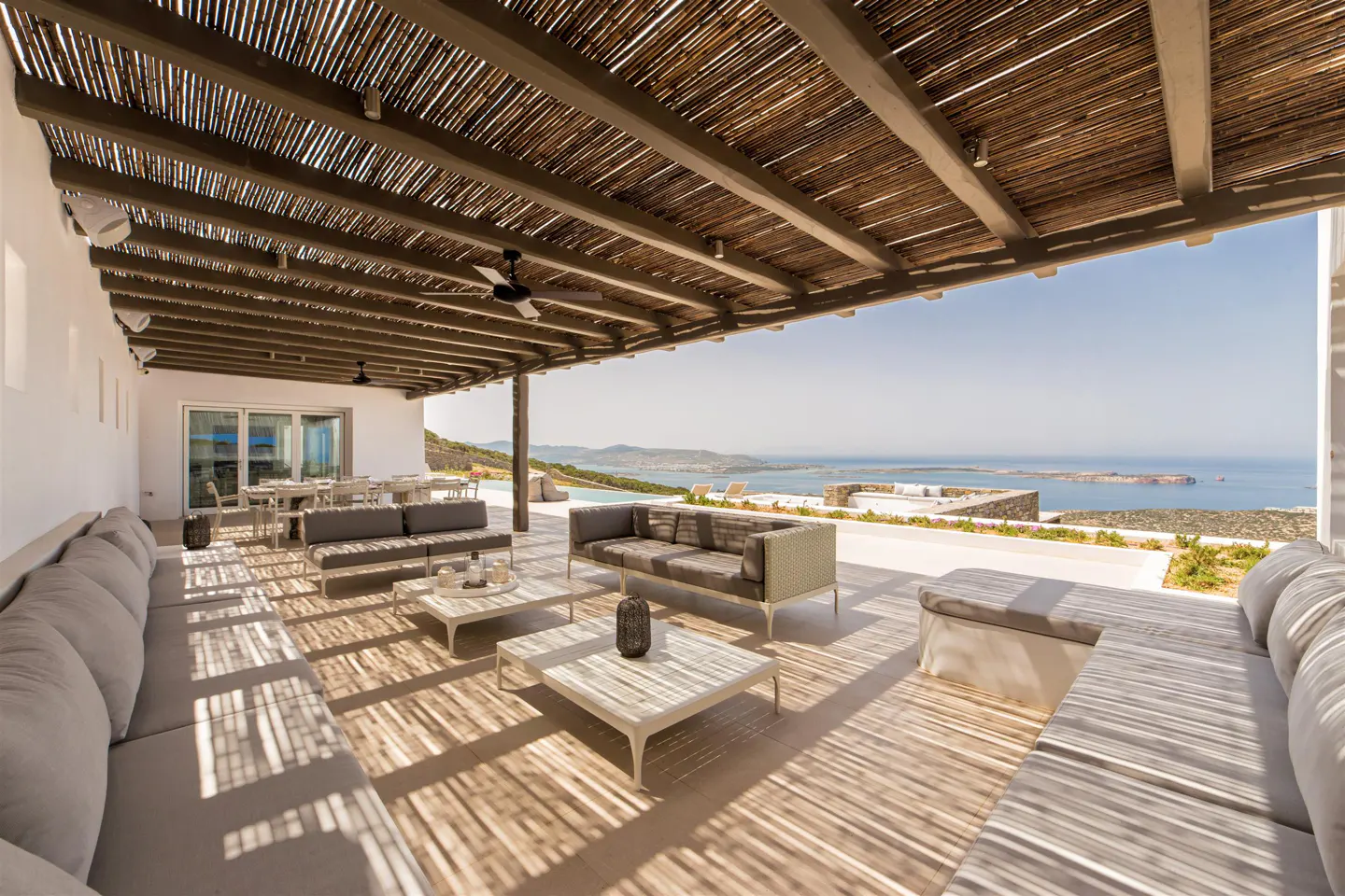 Covered outdoor patio with grey lounge seating, coffee tables, and a panoramic view of the sea and distant islands.