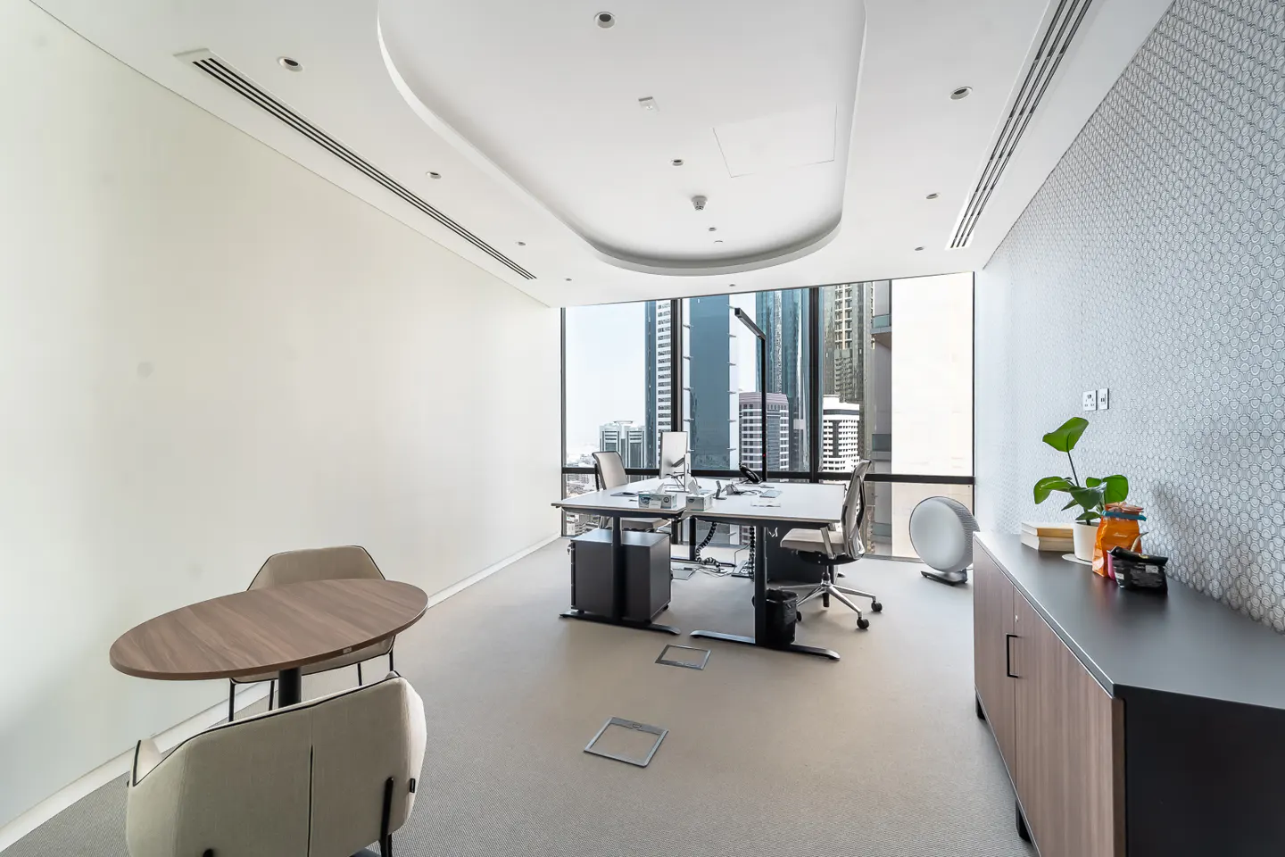 Bright office space with two desks, chairs, and a small round table. Large windows offer a city view. A cabinet with a plant sits against a patterned wall.