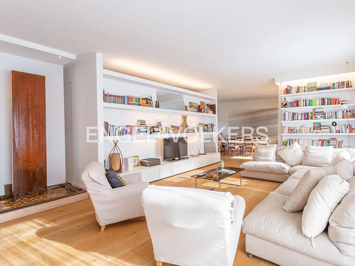 Bright living room with hardwood floors, white sofas, and built-in bookshelves filled with books. A modern water feature is on the left.