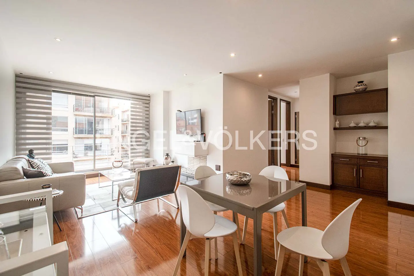 Bright, modern apartment interior with wood floors, a gray dining table with white chairs, and a living room with a large window.