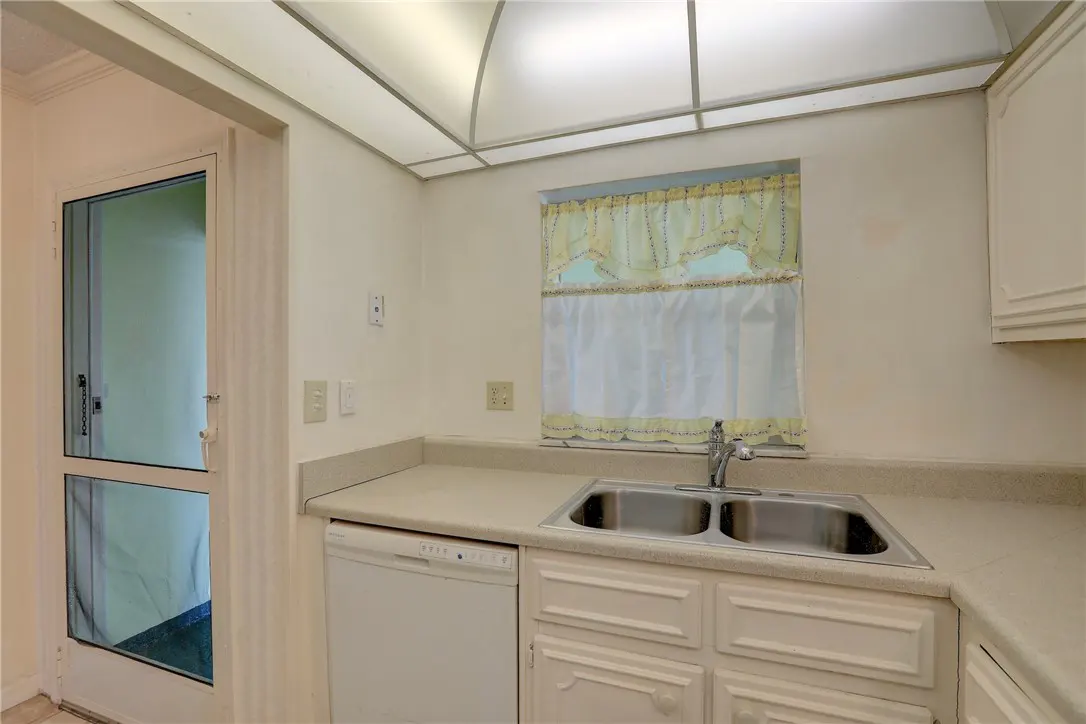A bright kitchen with white cabinets, a double sink, and a window with yellow curtains. A white dishwasher is to the left of the sink. A glass door is on the left.