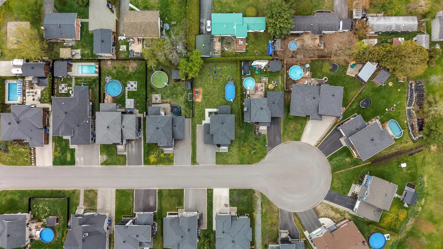 Aerial view of a suburban neighborhood with gray-roofed houses, green lawns, and a cul-de-sac street. Some houses have pools.