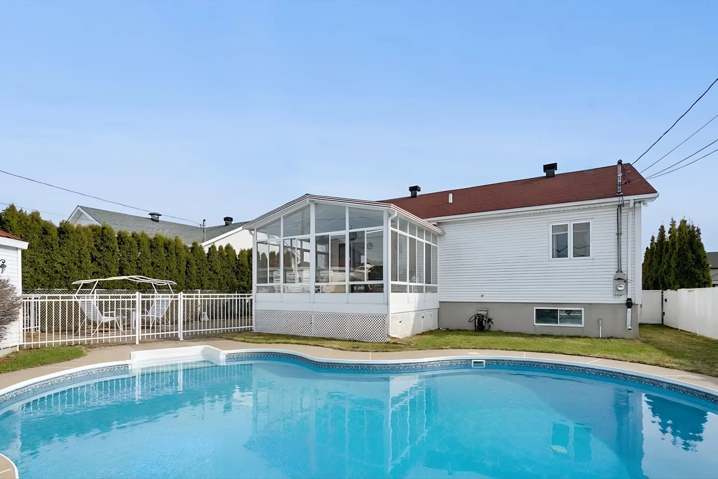 Backyard with a blue swimming pool, white house with a sunroom, and a white fence against a clear blue sky.