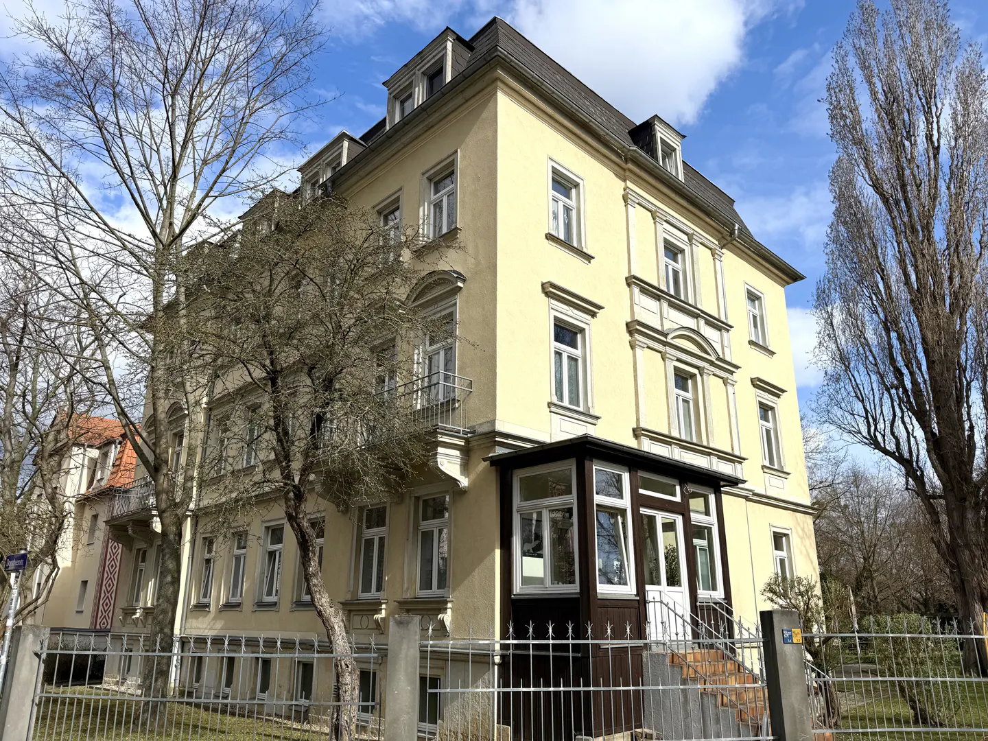 A three-story yellow building with white windows and a dark roof, framed by bare trees and a metal fence.