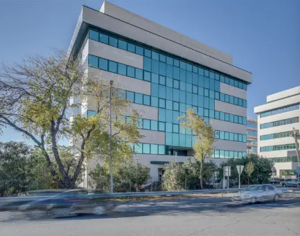 Modern office building with blue-tinted windows and a light gray facade, viewed from across a street with passing cars.