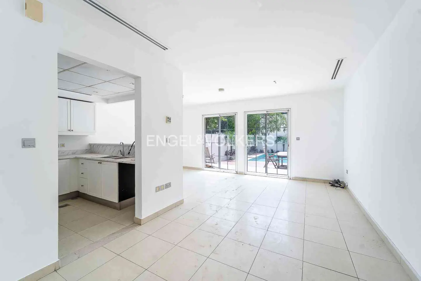Bright, empty room with white walls and tiled floor. Kitchen area visible through doorway. Sliding glass doors lead to a pool area.