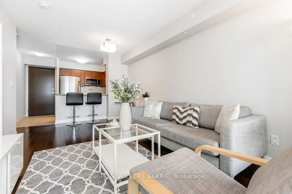 Bright living room with gray sofa, patterned rug, and glass coffee table. Kitchen with bar stools visible in the background.