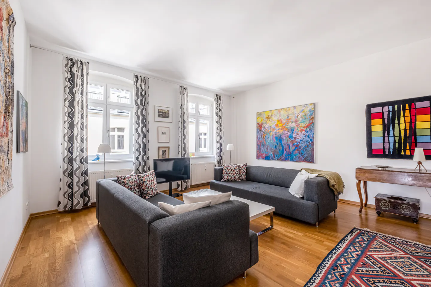 Bright living room with hardwood floors, two gray sofas, a white coffee table, and colorful artwork on the white walls. Windows with patterned curtains.
