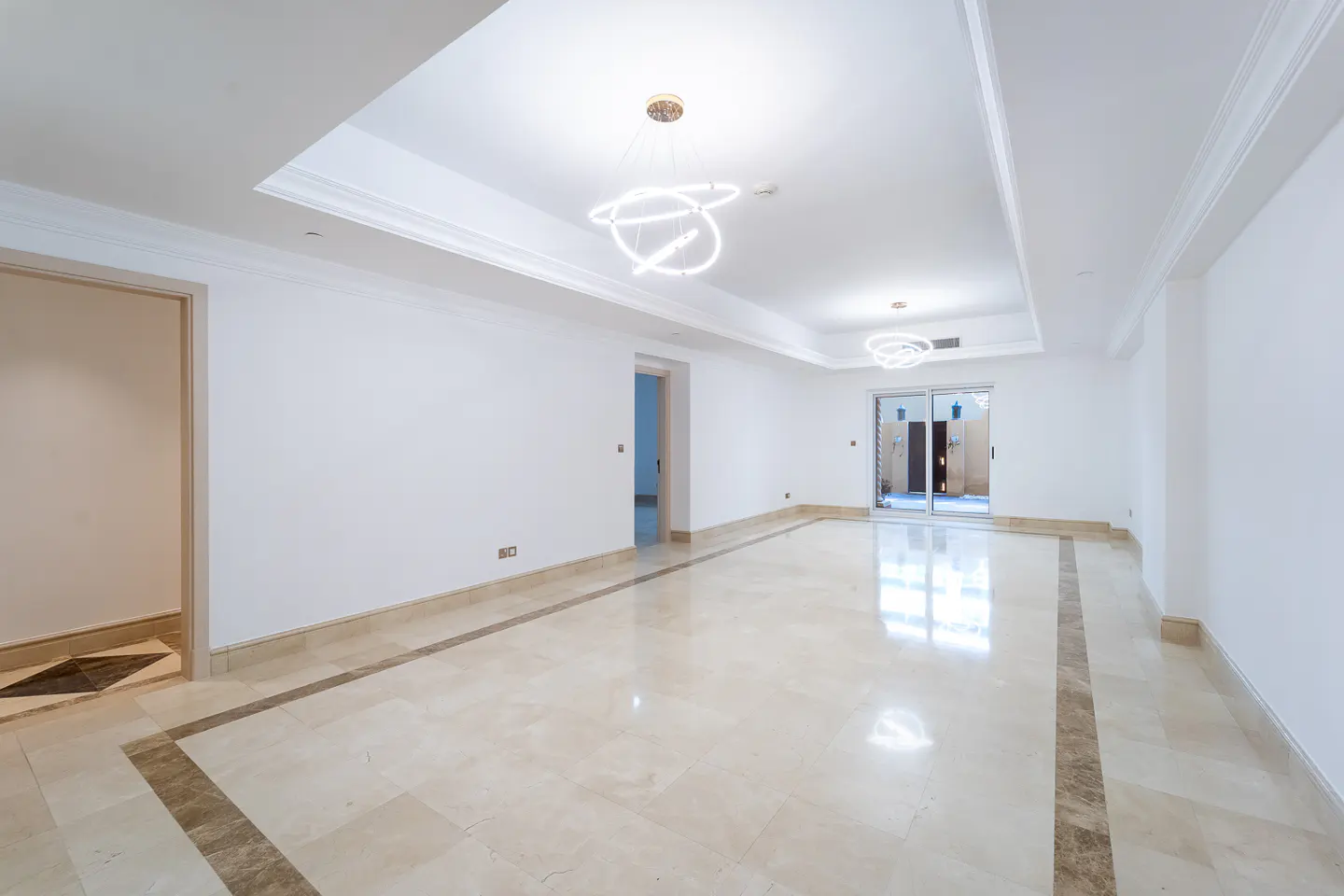 Bright, empty room with white walls, marble floors, and modern light fixtures. Sliding glass doors lead to an outdoor area.
