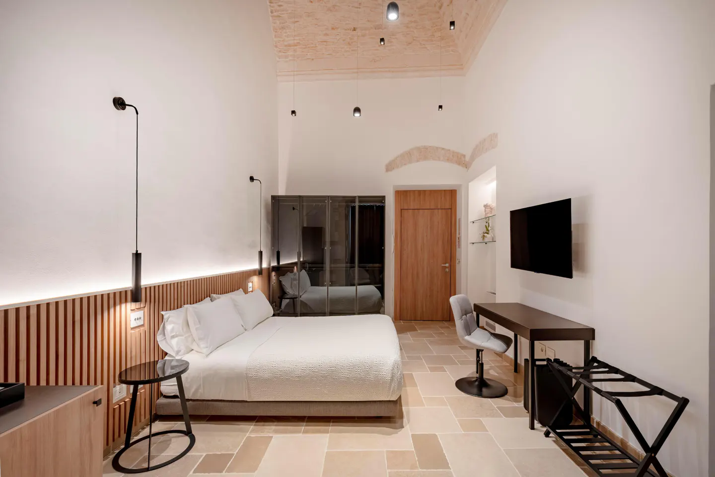 A modern bedroom with a white bed, desk, TV, and stone floors.