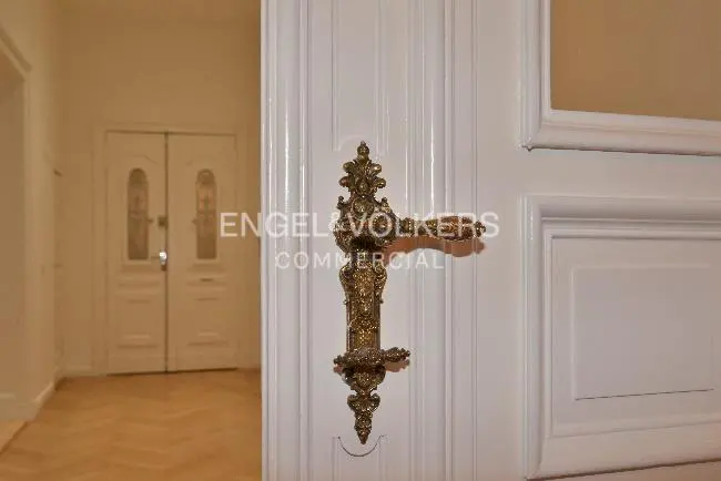Close-up of an ornate, gold handle on a white door, with a glimpse of a hallway and double doors in the background.