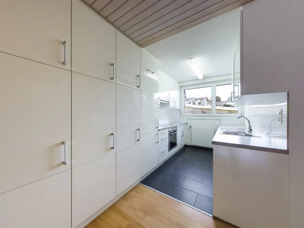 Modern galley kitchen with white glossy cabinets, dark tile flooring, light wood accents, and a window over the sink.