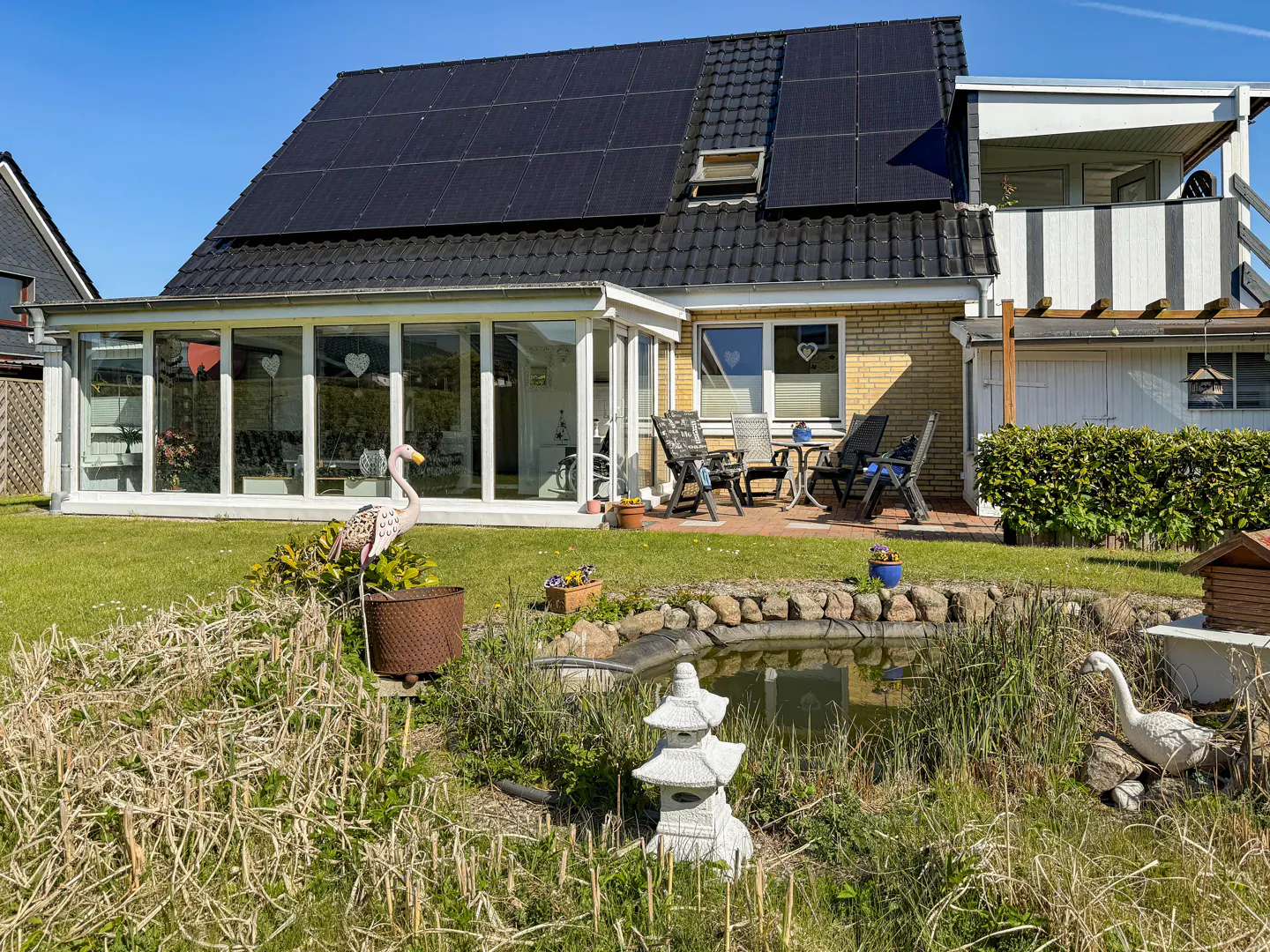 A house with solar panels, a sunroom, and a patio with chairs overlooks a garden with a pond and decorative statues.