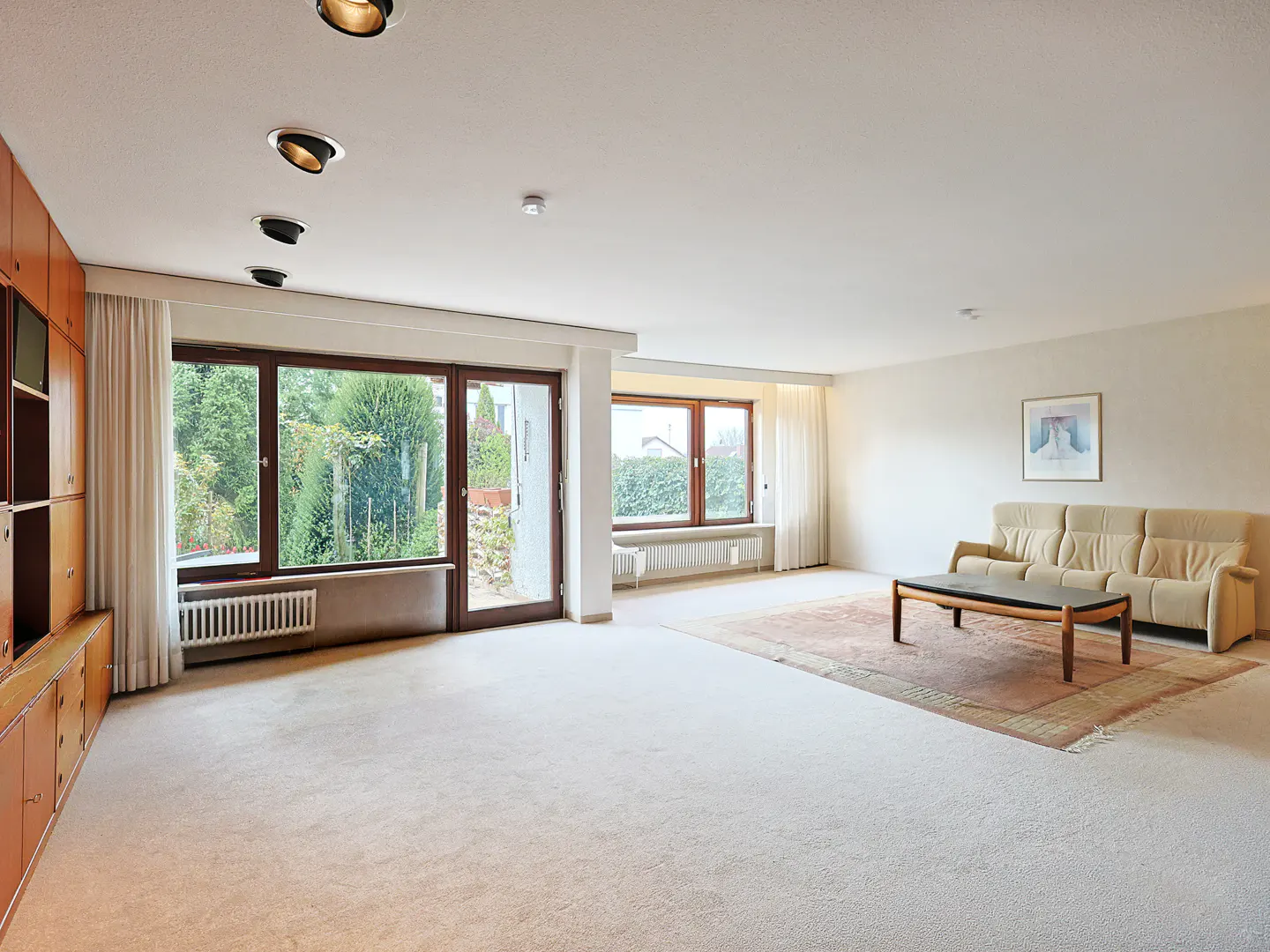 Bright living room with beige carpet, sofa, and wood coffee table on a patterned rug. Large windows offer a view of greenery.
