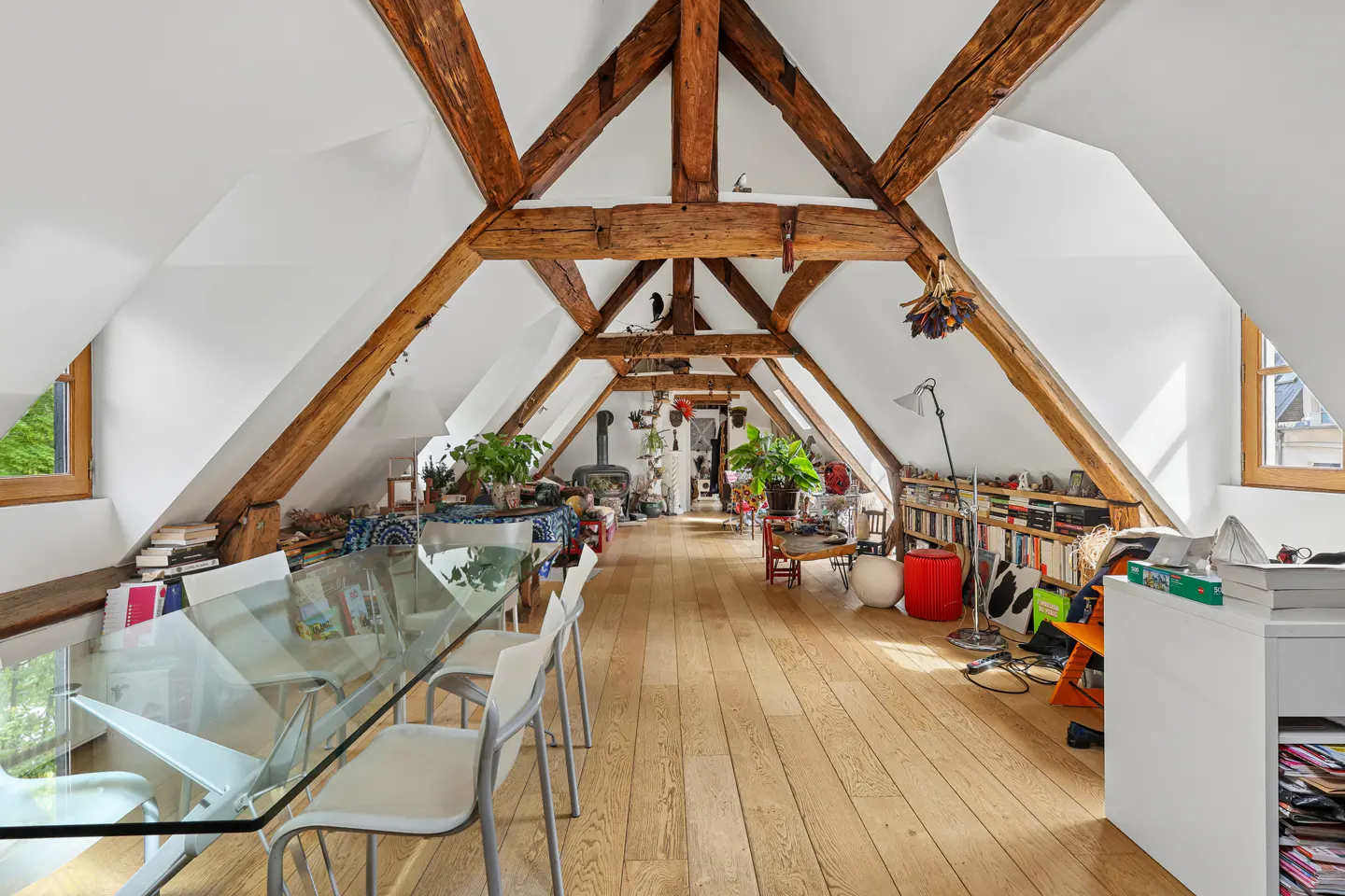 Attic room with exposed wooden beams, white walls, and light wood floors. A glass table and chairs are in the foreground.