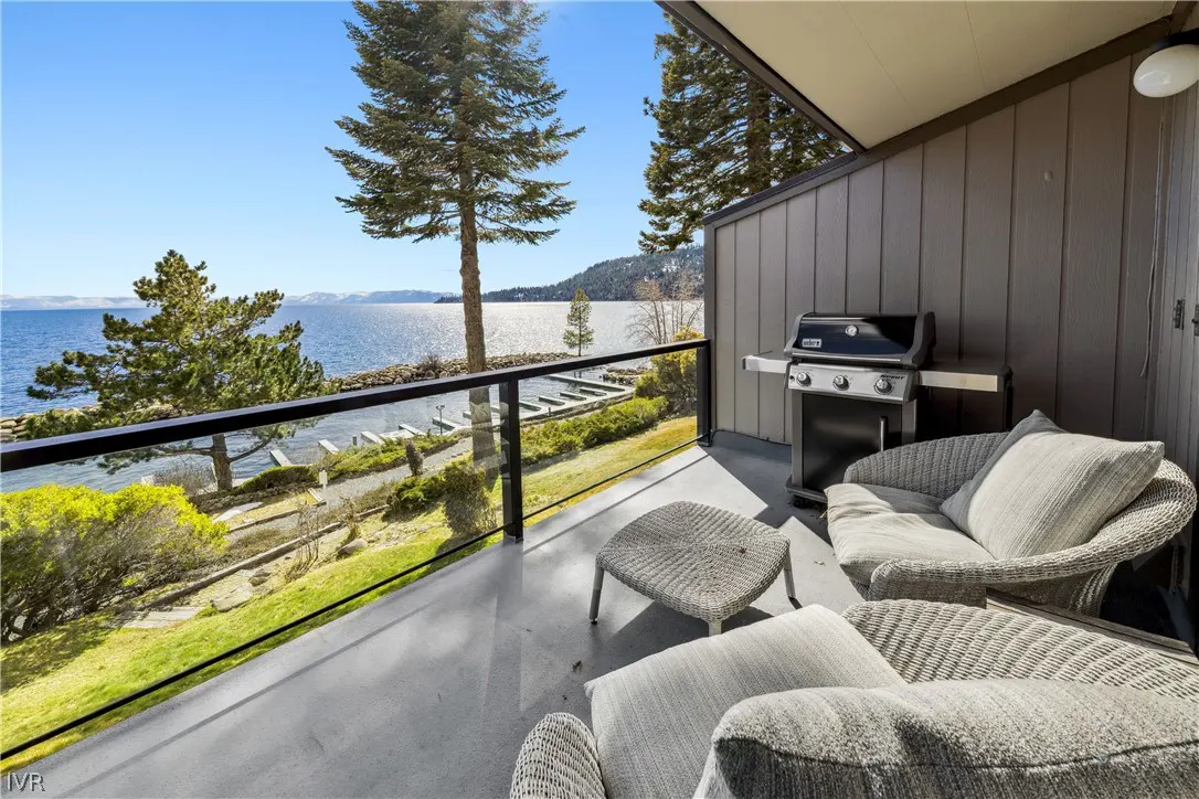 Balcony with wicker furniture and grill overlooking a lake. Trees and mountains are visible in the background under a blue sky.