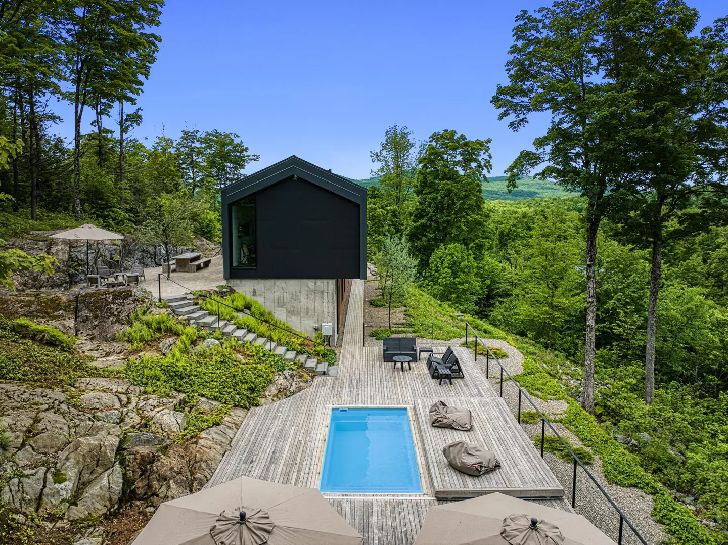 Modern black house with a pool on a wooden deck, surrounded by lush green trees and a clear blue sky.