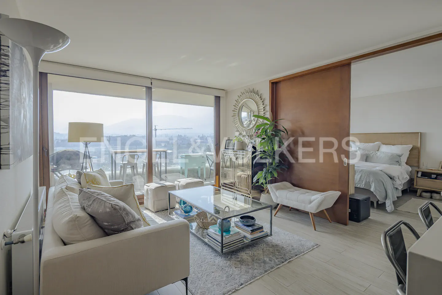 Bright apartment living room with white sofa, glass table, and balcony overlooking a city view. Bedroom visible through a sliding door.