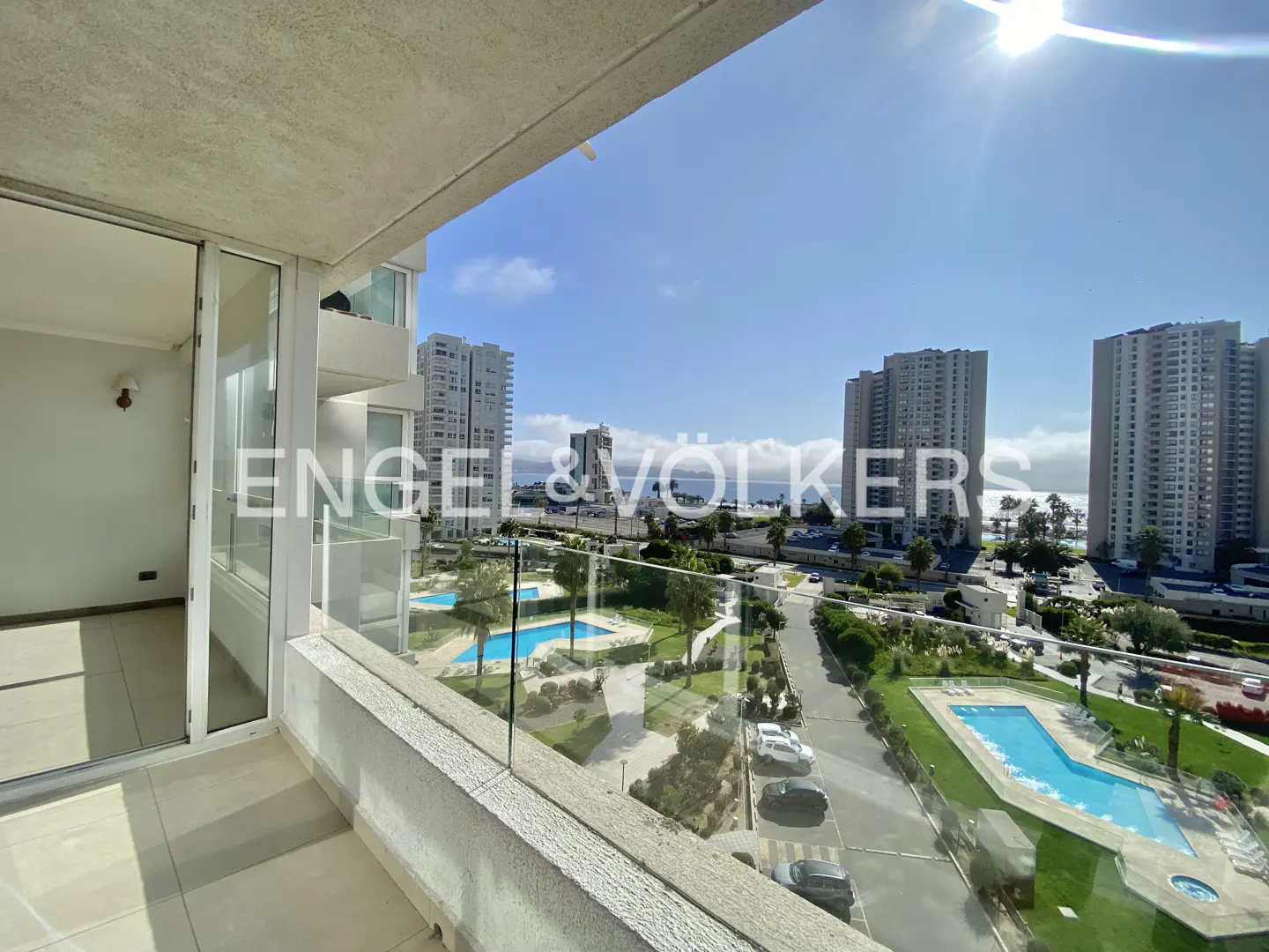 View from a balcony with glass railings overlooking pools, buildings, and the ocean on a sunny day.