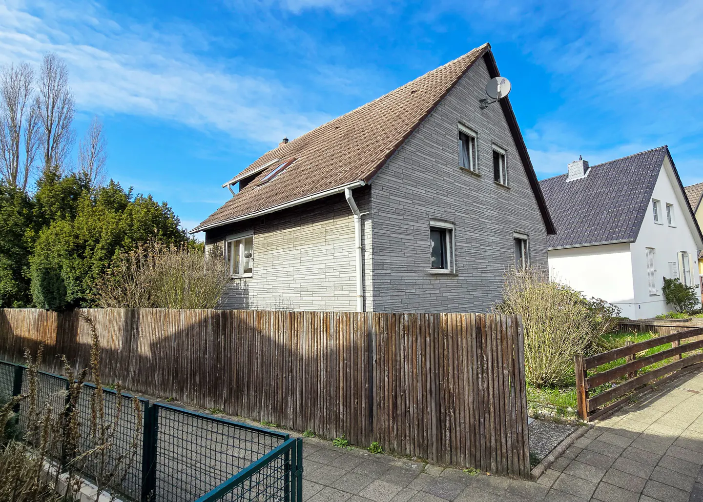 A two-story gray house with a brown roof and a wooden fence in front of it under a blue sky.