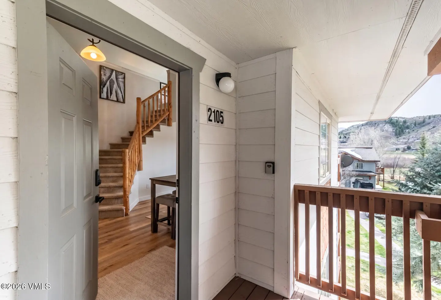 Open doorway to condo unit 2105. Stairs with wooden railing, table, and artwork visible inside. Balcony with brown railing overlooks a mountain view.