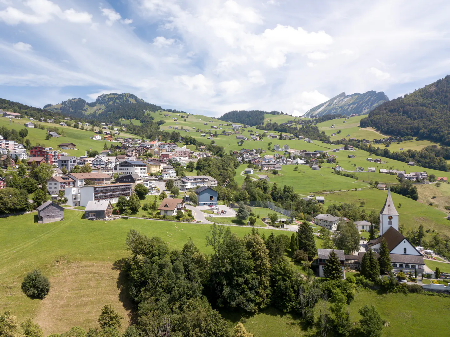 Scenic aerial view of a Swiss village nestled in green hills, featuring a church with a tall spire and scattered houses.