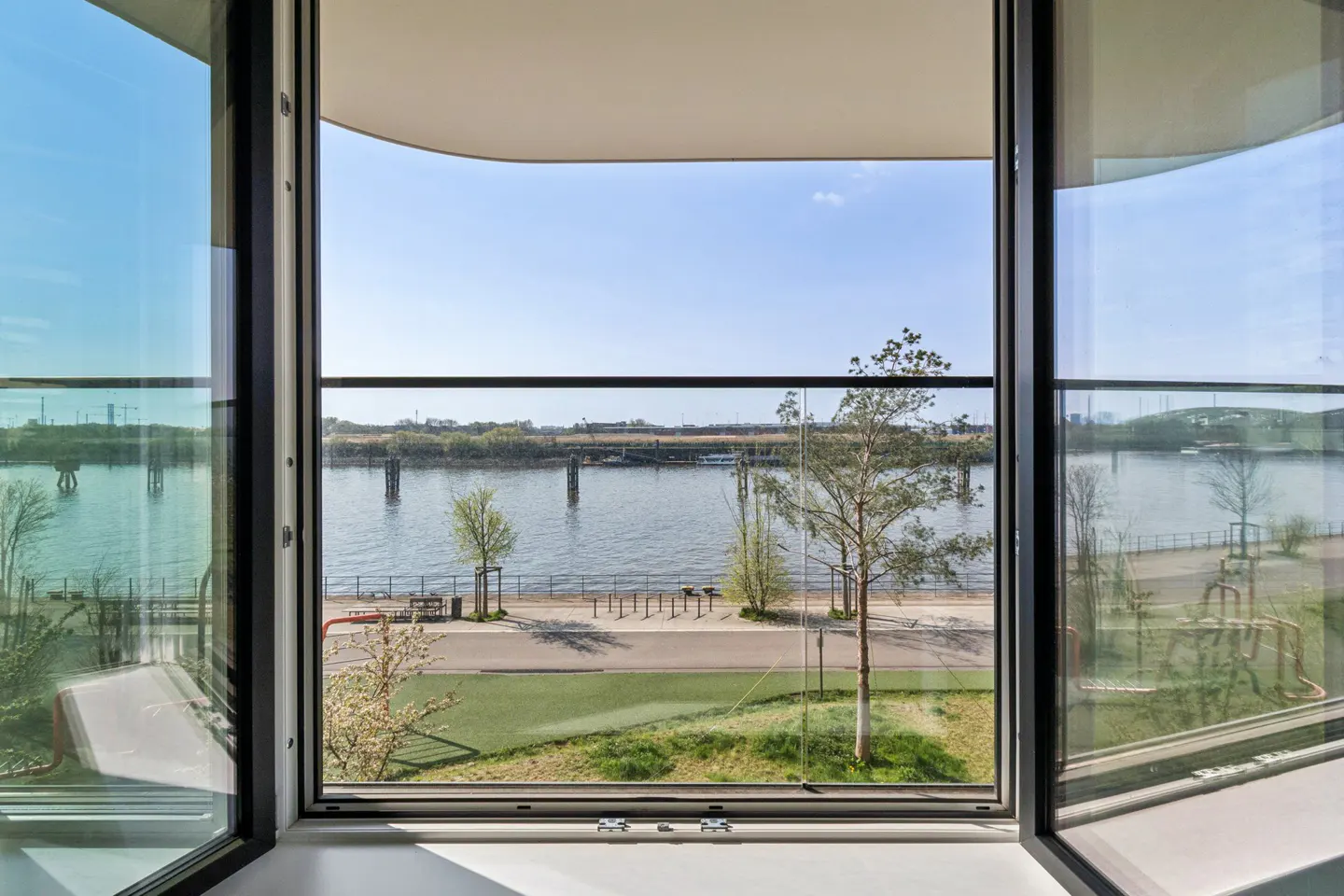 View from open window of river, trees, and walkway. Black window frame, blue sky, and green grass visible.