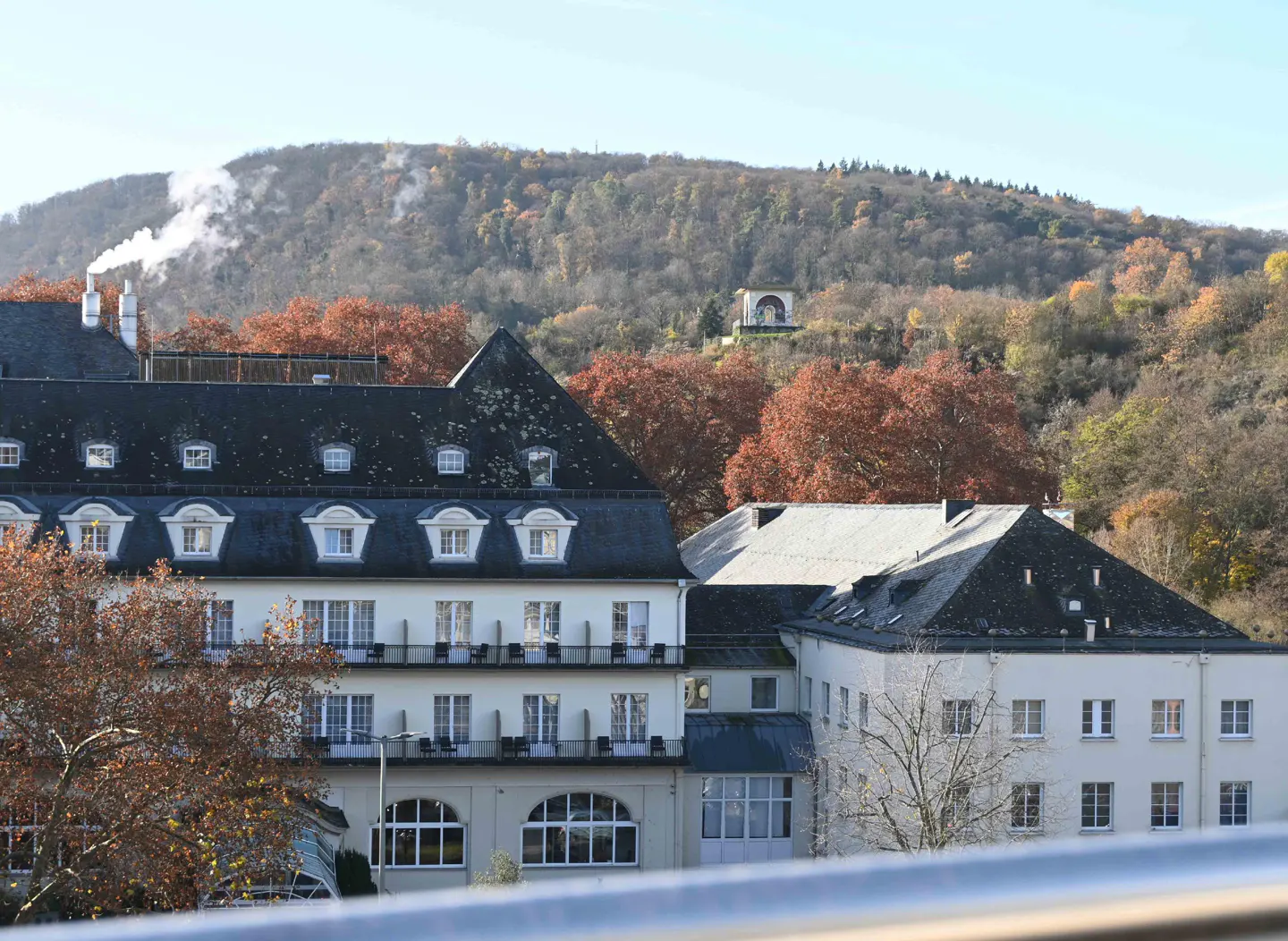 View of a white hotel with a dark roof, set against a backdrop of autumn trees and a distant hill.