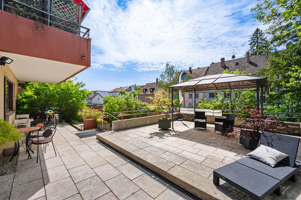 A stone patio with a gazebo, table, chairs, and lounge chair. Green trees and buildings are in the background.
