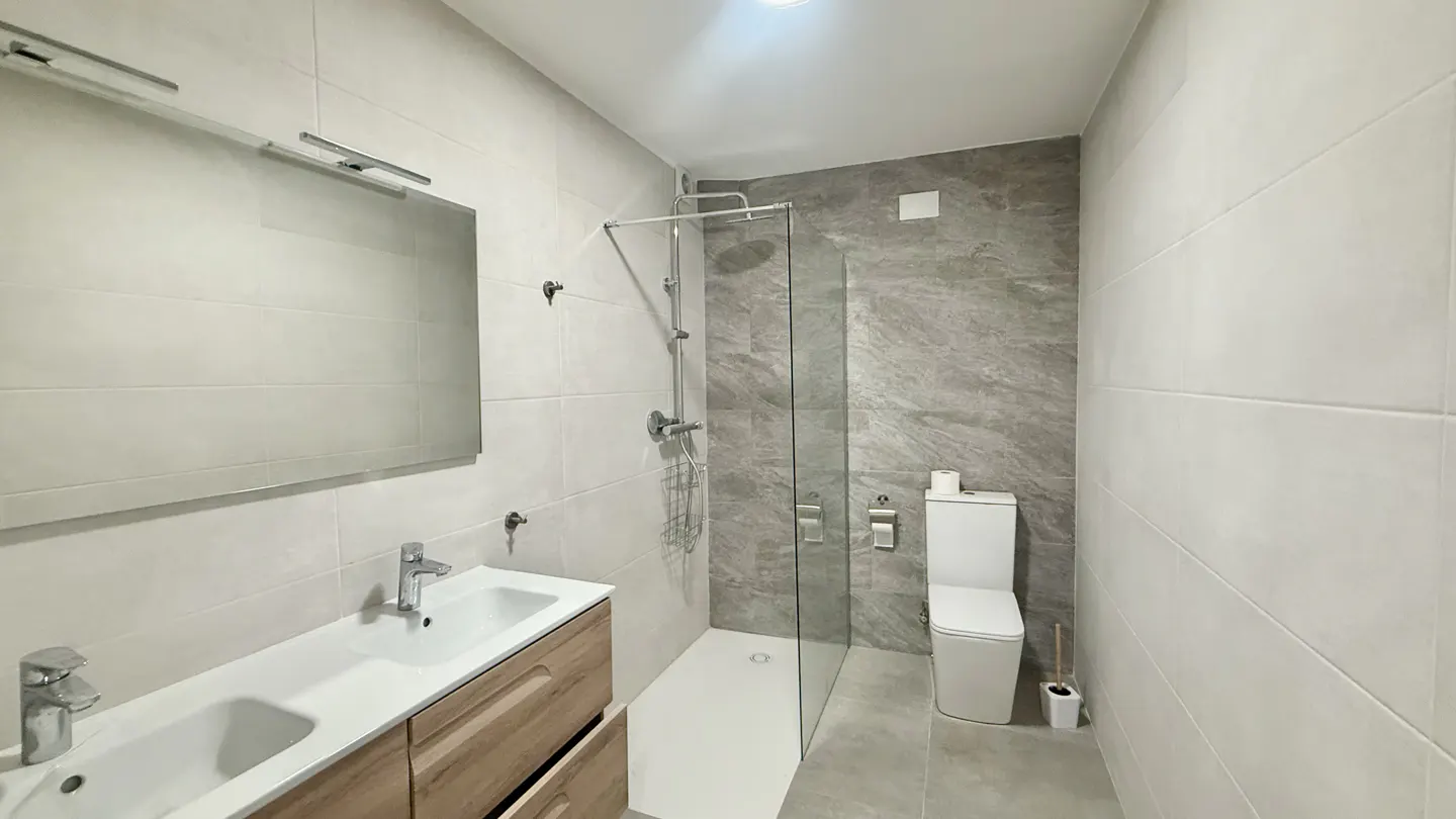 Bright bathroom with double sink vanity, large mirror, glass shower, and toilet against gray marble-look tile.