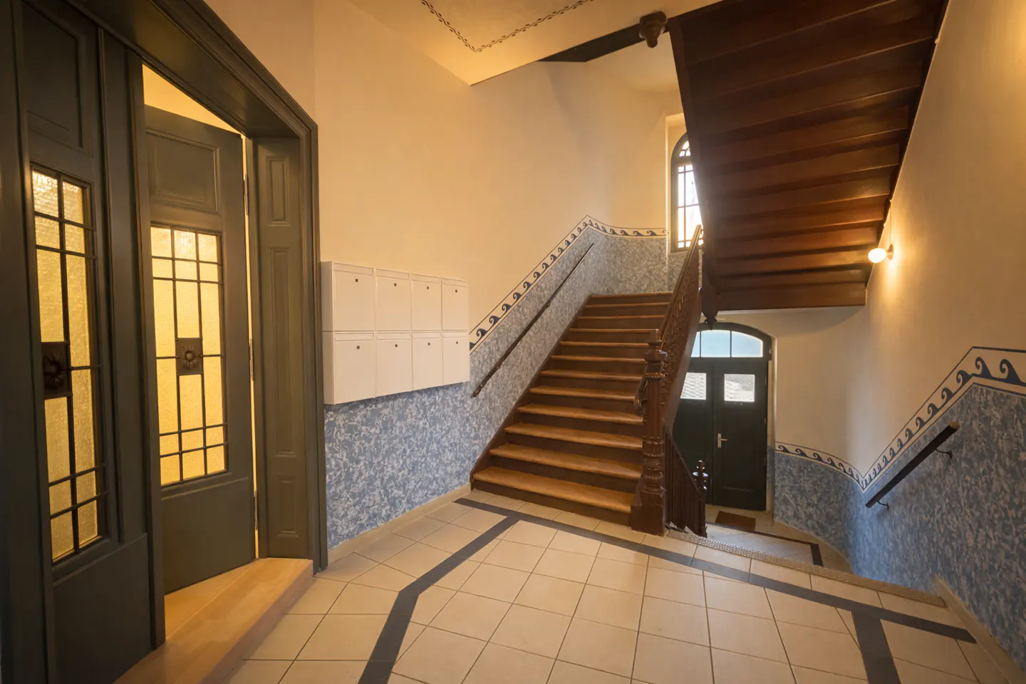 Interior view of a building's entrance hall with stairs. A blue door is open to the left. White mailboxes are on the wall. The stairs are wood with blue-patterned tile.