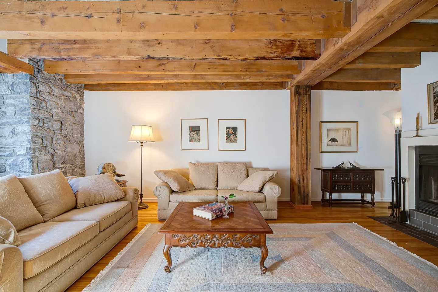 Living room with exposed wood beams, stone wall, and beige sofas. A patterned rug covers the hardwood floor.