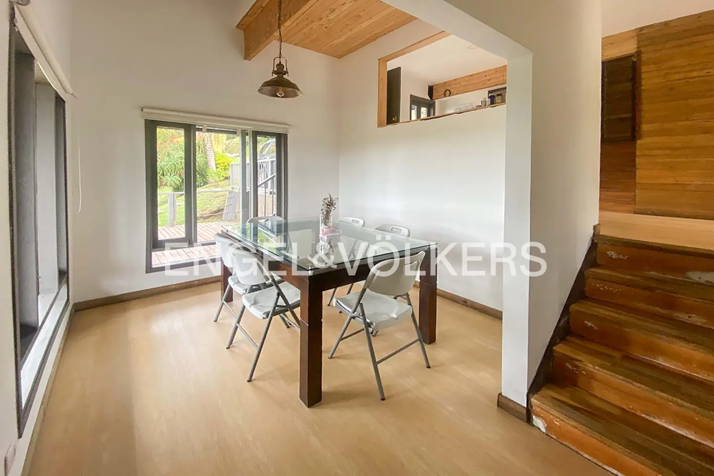 Bright dining room with a glass-top table, white folding chairs, and a view to the outside deck. Wooden stairs are on the right.