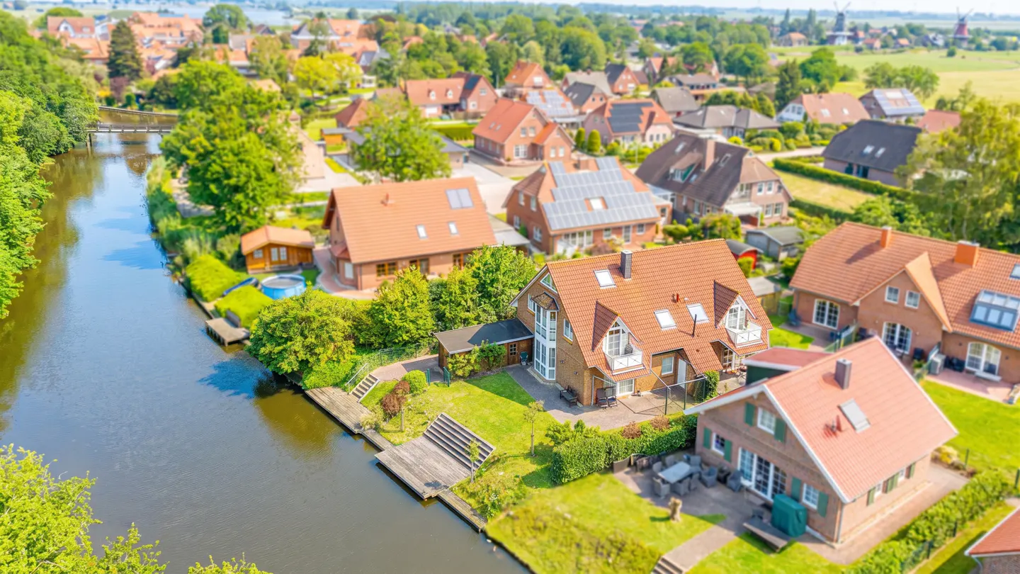 Aerial view of a quaint village with red-roofed houses along a canal, surrounded by lush green trees and fields.