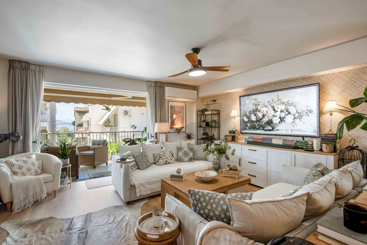 Bright living room with white sofas, wooden coffee table, large floral wall art, and a balcony with outdoor seating.