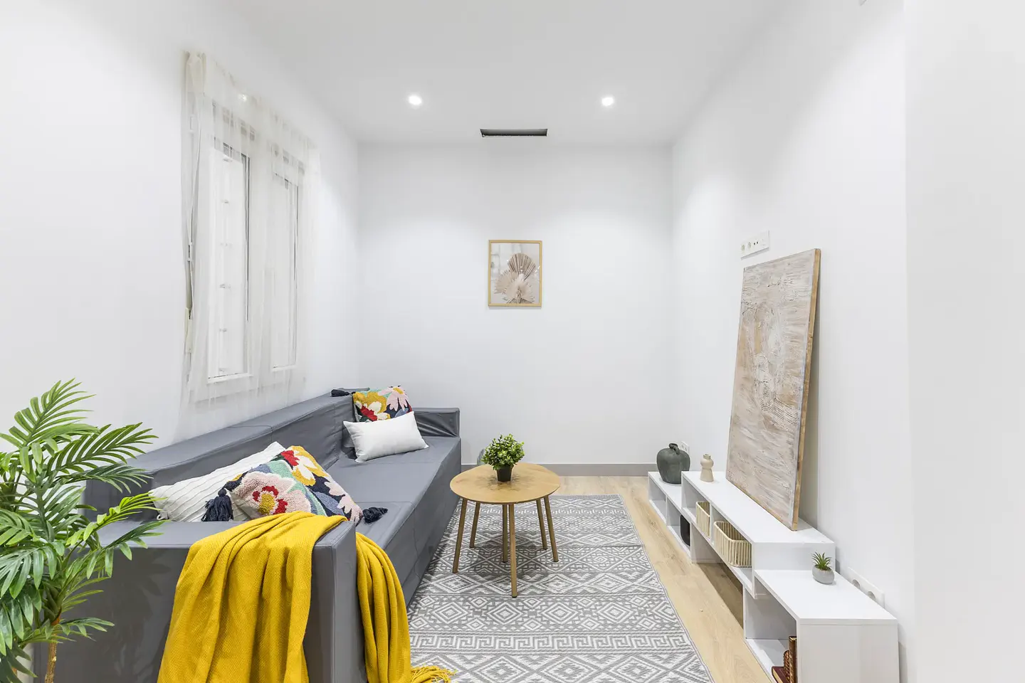 Bright living room with gray sofa, yellow throw, patterned pillows, and a round wood table on a gray patterned rug. White walls and light wood floors.