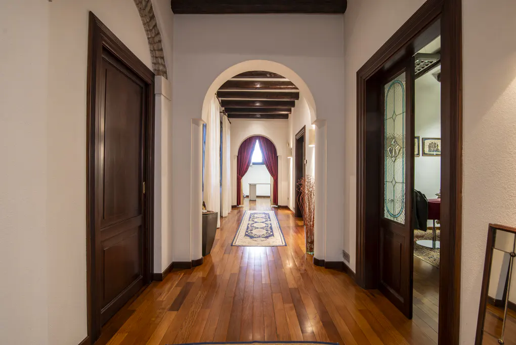 Hallway with wood floors, white walls, and dark wood doors. An arched doorway leads to a room with red curtains. A blue patterned rug runs down the center.