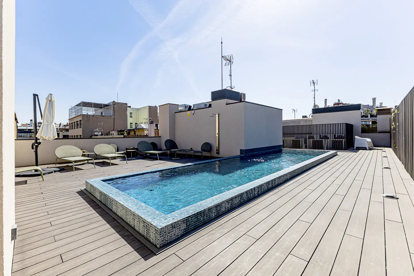 Rooftop pool with lounge chairs on a sunny day. The pool has blue tiles and the deck is made of light brown wood.